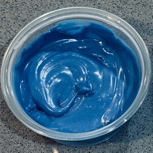 May include: A transparent plastic pot filled with a vivid blue, creamy substance. The contents have a smooth, glossy texture, with visible swirls and peaks. The pot sits on a speckled grey surface.