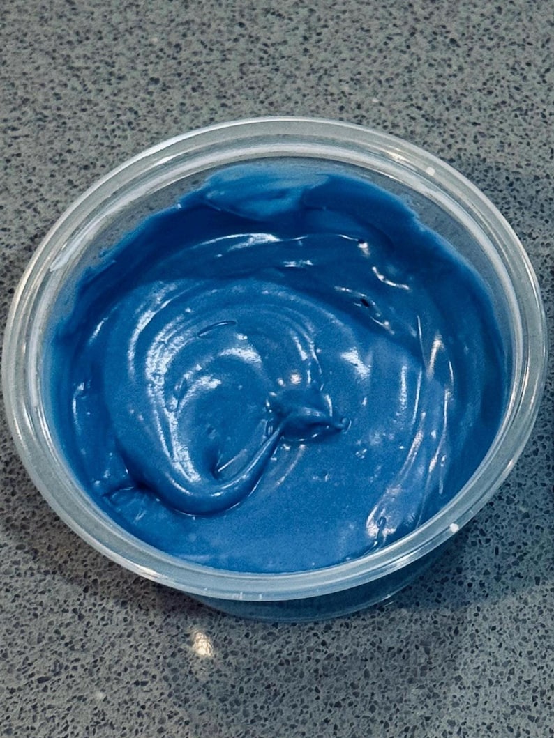 May include: A transparent plastic pot filled with a vivid blue, creamy substance. The contents have a smooth, glossy texture, with visible swirls and peaks. The pot sits on a speckled grey surface.