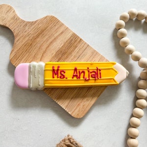 May include: A yellow and pink sugar cookie shaped like a pencil with the words "Ms. Anjali" written in red icing.