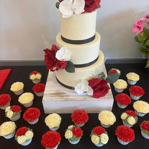 K&ouml;nnte beinhalten: Eine dreist&ouml;ckige wei&szlig;e Hochzeitstorte, verziert mit schwarzem Band und roten und wei&szlig;en Blumen. Die Torte ist von Cupcakes mit rotem und wei&szlig;em Frosting umgeben.