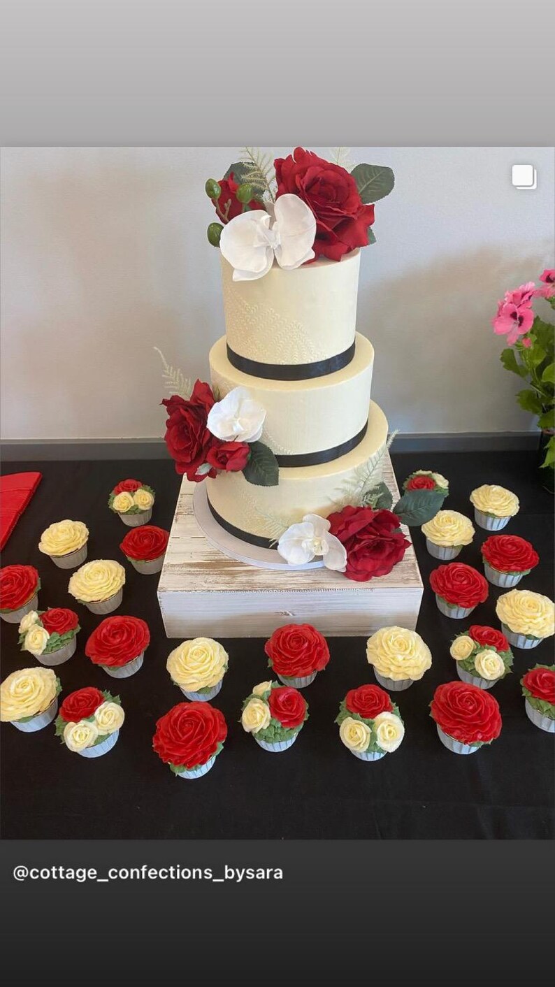 K&ouml;nnte beinhalten: Eine dreist&ouml;ckige wei&szlig;e Hochzeitstorte, verziert mit schwarzem Band und roten und wei&szlig;en Blumen. Die Torte ist von Cupcakes mit rotem und wei&szlig;em Frosting umgeben.