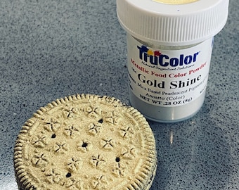 May include: A gold-coloured, decorated biscuit next to a jar of TruColor Gold Shine metallic food colour powder. The jar has a white lid with the words "GOLD SHINE" printed on it. The biscuit has a detailed starburst pattern.
