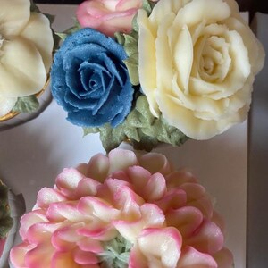 May include: Close-up of cupcakes decorated with buttercream flowers. The cupcakes feature a variety of colours, including pink, blue, and yellow. The flowers are intricately detailed, resembling roses and other blooms. The text "@cottage_confections_bysara" is visible.