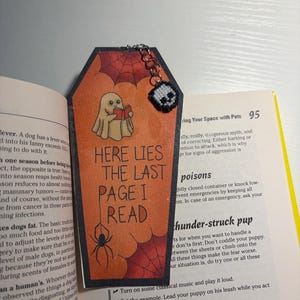 May include: A coffin-shaped bookmark with an orange background, featuring a ghost reading a red book, a black spider, and red spiderwebs. The text reads "HERE LIES THE LAST PAGE I READ". A small, black and white skull charm hangs from a silver chain.