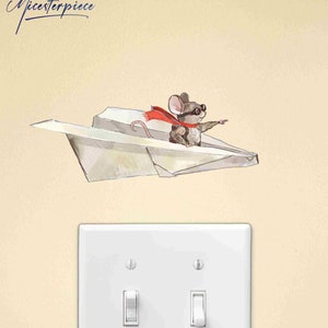 May include: A white paper airplane with a small mouse wearing aviator goggles and a red scarf flying in the air. The mouse is pointing with its right paw. The image is on a light beige wall with a white light switch.