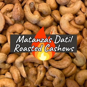 May include: Close-up of a pile of roasted cashews. The cashews are a light brown color and have a slightly salty and sweet flavor. The text "Matanzas Datil Roasted Cashews" is printed on a black banner with a red flame graphic.