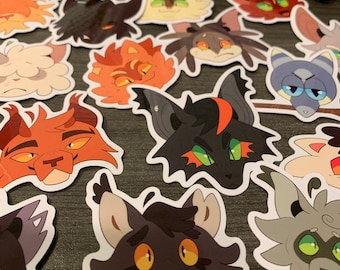 May include: A collection of colorful cat-themed stickers. The stickers feature various cat faces with different expressions and colors, including orange, black, gray, and cream. The stickers are die-cut and have a white border.