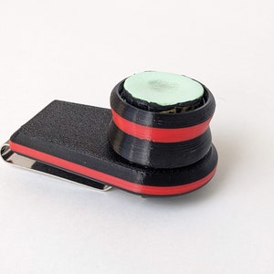 Könnte beinhalten: Ein schwarz-roter Geldclip mit strukturierter Oberfläche und silbernem Clip. Ein schwarz-rotes Objekt mit hellgrüner Oberseite ist am Geldclip befestigt. Das Objekt hat eine zylindrische Form.