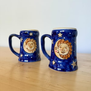 May include: Two blue ceramic mugs with handles, decorated with a sun and moon design. The sun has a smiling face and is surrounded by a crescent moon and stars. The mugs are on a wooden surface.