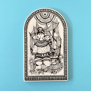 Peut inclure: Plaque décorative en céramique noire et blanche avec un dessus en forme d'arche. La plaque présente une illustration détaillée d'une personne tenant un enfant, un chat et le texte "NORTH NOR NOR". La bordure est décorée d'un motif pointillé.