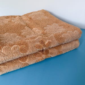 May include: Two folded, light brown bath towels. The towels have a floral pattern along the edges. The towels are made of a soft, absorbent material. The towels are stacked on top of each other on a blue surface.