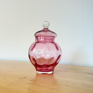 May include: A pink glass jar with a lid. The jar has a round body with oval indentations around the sides. The lid is a smaller version of the jar's body, with a clear glass knob on top. The jar is on a wooden surface.