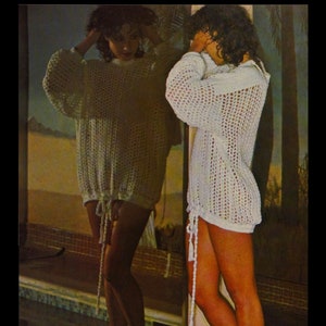 Puede incluir: Una mujer lleva un suéter de crochet blanco de corte holgado y diseño largo y fluido. El suéter tiene un delicado patrón de calado y un detalle de borla en la parte inferior. La mujer está de pie frente a un espejo, y su reflejo es visible en el fondo.