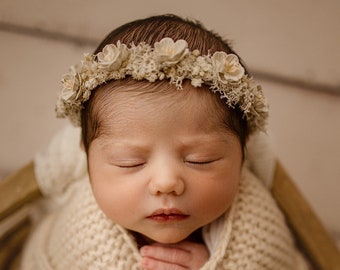 May include: A newborn baby girl sleeping peacefully in a cream-colored knitted wrap. She is wearing a flower crown with white blossoms.