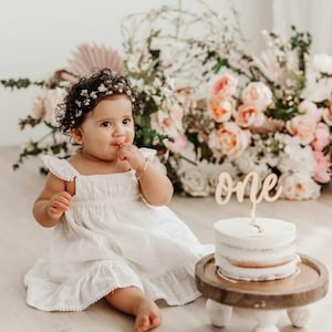 May include: A baby girl wearing a white dress and a flower crown sits on a white floor in front of a floral backdrop. She is eating cake with the word "one" on top.