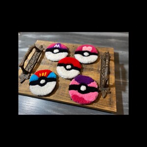 Puede incluir: Cinco posavasos redondos y tupidos de colores que se asemejan a Poké Balls, cada uno con un diseño único. Los posavasos están dispuestos sobre una bandeja de madera con asas de metal ornamentadas. Los posavasos presentan varios colores, incluyendo rojo, rosa, azul, morado y blanco.