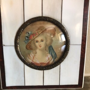 May include: A framed miniature portrait of a woman wearing a straw hat with a blue ribbon. The portrait is set against a white background and framed in a dark wood frame.