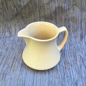 May include: A cream-colored ceramic pitcher with a rounded body, a curved handle, and a pouring spout. The pitcher has a smooth, glossy finish and is likely used for serving liquids. The design is simple and classic, suitable for various kitchen styles.
