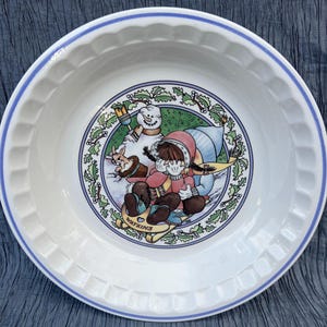 May include: White ceramic pie dish with a blue rim and a scalloped edge. The center features a winter scene with a snowman, a child on a sled, and a rabbit. The sled has the word "WATKINS" on it.