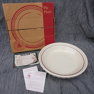 May include: A cream-colored ceramic pie plate with a thin maroon rim, accompanied by its original cardboard box labeled Pie Plate, a small informational booklet, and a product card detailing Longaberger Pottery and Traditional Red.