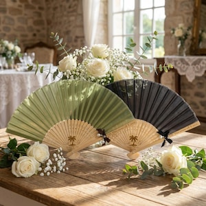 以下が含まれることがあります： 緑、黒、天然木色の3つの装飾的な扇子が、白いバラと緑の葉とともに木製の表面に配置されています。扇子にはヤシの木の模様と「All Because of Love」の文字が描かれています。