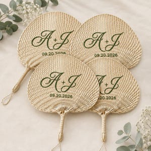 Puede incluir: Cuatro abanicos de mano tejidos de hoja de palma con una textura natural de color beige claro. Cada abanico presenta letras bordadas en cursiva de color verde oscuro que muestran las iniciales A + J y la fecha 09.20.2026. Forma redondeada.