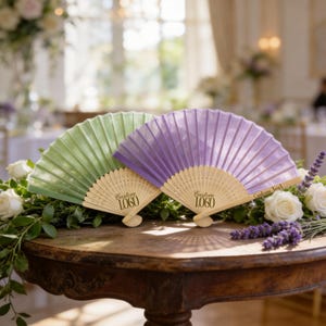 May include: Two open hand fans, one light green and one purple, rest on a wooden table. The fans have wooden handles and are decorated with the text "Custom Logo 1060". White roses and lavender sprigs are arranged around the fans.