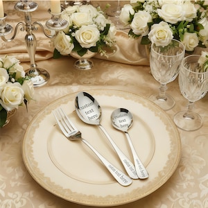 May include: A formal table setting with a cream-colored plate, silverware, and crystal glasses. The silverware includes a fork, a spoon, and a smaller spoon, all engraved with "custom text here." The table is decorated with white roses and a silver candelabra.
