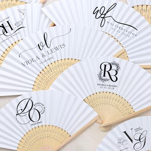 May include: A collection of white paper fans with wooden handles, each featuring custom black text and dates. The fans are folded and fanned out, displaying various names and dates, ideal for weddings or special events.