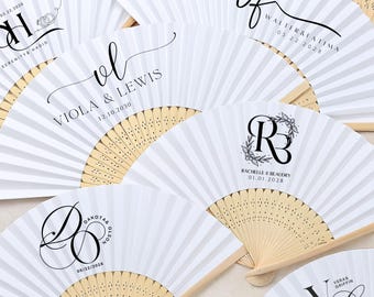 May include: Several white paper fans with wooden handles, each printed with custom black text and dates. The fans are folded and fanned out, displaying various names and dates, suitable for weddings or special events.