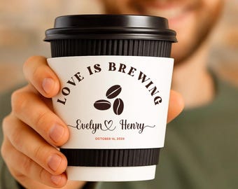 May include: A black coffee cup with a white sleeve that reads "LOVE IS BREWING" with coffee bean graphics. The sleeve also includes the names "Evelyn & Henry" and the date "OCTOBER 14, 2026".