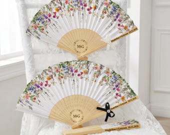 May include: Three white folding fans with floral designs and wooden handles. The fans feature a delicate pattern of colorful flowers and the initials "M&G" within a wreath. The text "Madeline + Grayson 10.22.2028" is also visible.