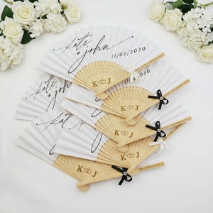 Op de afbeelding: Witte papieren waaiers met houten handvatten, gepersonaliseerd met de namen "Kate + John" en een datum. De houten handvatten zijn gegraveerd met "K J" en een ringontwerp. Sommige waaiers hebben zwarte of witte linten.