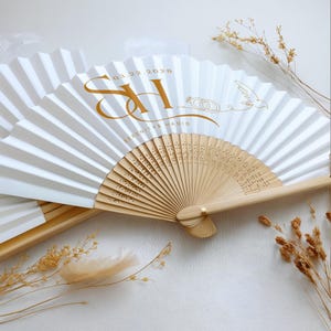May include: White paper folding fan with a light brown bamboo handle. The fan has gold lettering with the date 03.22.2028, initials, and the names Serenity & Hadid. A dove and wedding rings are also featured.