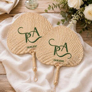 May include: Two round, woven palm leaf hand fans with a natural tan texture. Each fan features an embroidered dark green monogram of the letters P and A in a cursive script, with the date 09.16.2026 stitched in green below the monogram.