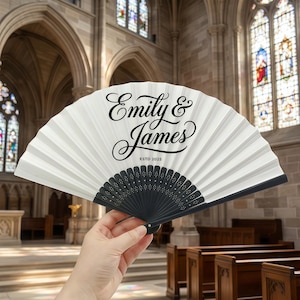 May include: A white folding fan with black detailing. The fan is inscribed with the names "Emily & James" in a decorative script, and "ESTD 2025". The fan is held in a church setting, possibly for a wedding or special occasion.