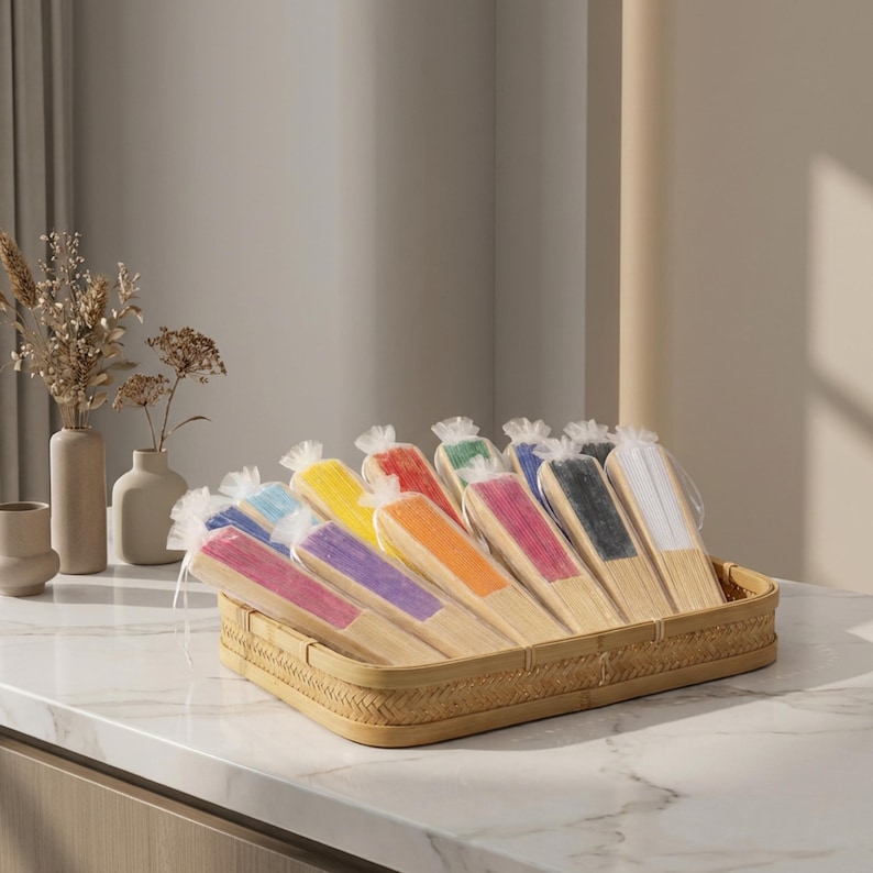 Op de afbeelding: Een verzameling handwaaiers met houten frames en stof in diverse kleuren, waaronder roze, paars, blauw, geel, oranje, rood, groen, zwart en wit, elk individueel verpakt in doorzichtig plastic en gerangschikt in een gevlochten mand.