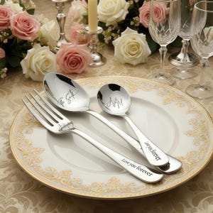 May include: A set of engraved cutlery, including a fork, spoon, and small spoon, arranged on a white plate with a gold rim. The spoon and small spoon have initials engraved. The fork and spoon have the words "Love you forever" engraved. Floral arrangements and glassware are in the background.