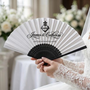 May include: A white folding fan with black detailing, held open. The fan displays the names "James & Catherine" and the phrase "For God so loved the world" in black script. A heart and cross design is at the top.