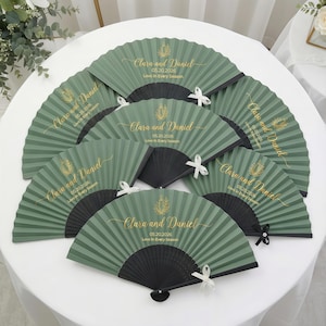 Puede incluir: Abanicos de papel verde con mangos negros, dispuestos sobre una superficie blanca. Cada abanico presenta texto dorado: "Clara and Daniel 05.20.2026 Love in Every Season" y un lazo decorativo.