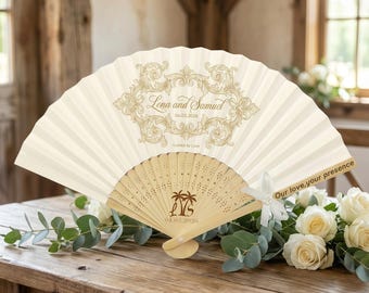 Op de afbeelding: Een crèmekleurige vouwventilator met een decoratief goudkleurig ontwerp en de namen "Lena en Samuel" en de datum "04.05.2026". De ventilator is gemaakt van papier en hout, met een gesneden houten handvat. De tekst "Our love, your presence" staat op de ventilator.