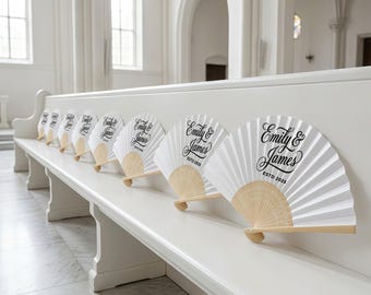 May include: A row of white paper fans with wooden handles, each bearing the text "Emily & James ESTD 2025" in black script. The fans are arranged on a white bench inside a bright, white-walled building, likely a church or event space.