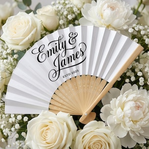 May include: A white paper fan with a wooden handle, featuring the names "Emily & James" and "ESTD 2025" in black script. The fan is surrounded by white roses and peonies, creating a wedding theme.