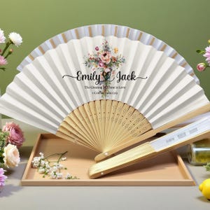 May include: Two white paper fans with a floral design and the names "Emily & Jack" printed on them. The text "The Greatest of these is Love" and "1 Corinthians 13:13" are also printed on the fan. The fans are on a wooden tray.