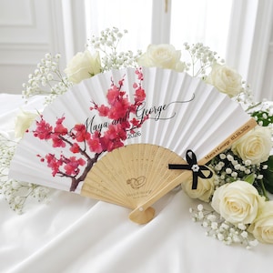 Pode incluir: Um leque branco dobrável com um design floral e os nomes "Maya and George" impressos em escrita preta. O leque apresenta um ramo floral vermelho e uma fita preta. A data 02.10.2026 também está impressa.