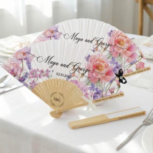 May include: Two white paper fans with floral designs and the names "Maya and George" printed on them. The fans have a wooden handle and are decorated with pink and purple flowers. The date "02.10.2026" is also printed on the fan.