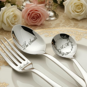 May include: A set of polished silverware, including a fork and two spoons, arranged on a white plate. The fork and one spoon are engraved with "The Smith." The other spoon features the initials "A M" within a wreath design. Soft pink and white roses are in the background.