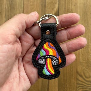 May include: Black leather keychain with a colorful embroidered mushroom design. The mushroom features a rainbow pattern on the cap and a white underside. The keychain has a silver metal ring.