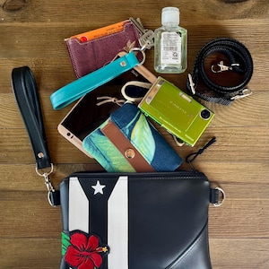 May include: A black clutch with a Puerto Rican flag design and a red hibiscus flower. The image also shows a teal wrist strap, keys, a small green camera, a phone, a card holder, a bottle of hand sanitizer, and a black camera strap.