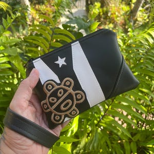 May include: Black and white clutch with a star and stripe design, featuring a gold-colored Taíno sun symbol. The clutch has a wrist strap and a zipper closure. The background is a lush green foliage.